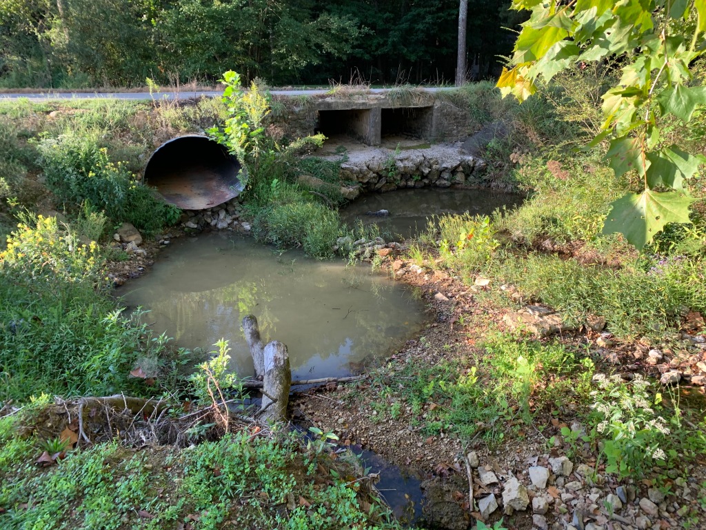 Shallow, dirty puddles sit in a creek bad next to a road. Three culverts are visible underneath the road, but all three are above the water level.