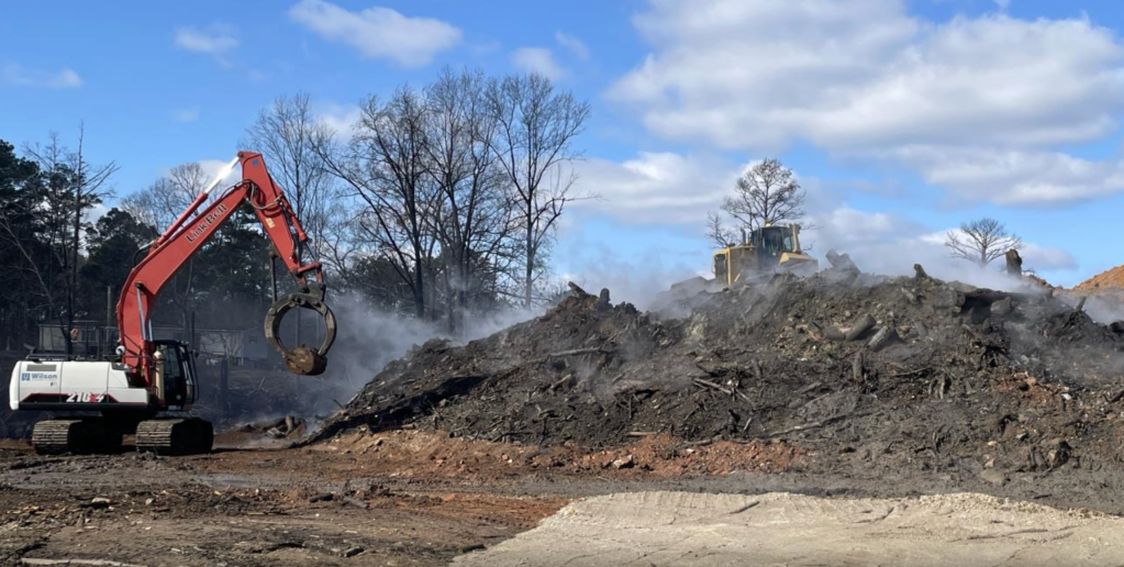 A large pile of smoking black soil takes up most of the image. A machine picks up what looks like a metal barrel on the left side of the pile, and a tractor is on top covering the pile with dirt to smother the fire.