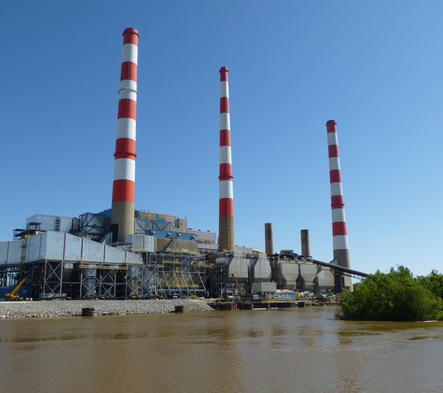 An electric power plant with three red-and-white-striped smokestacks, sits on the bank of Mobile River, which is brown and cloudy.