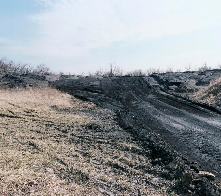 From the right side of the image, a road of black ash travels toward a pile of ash in the background, with tracks from large vehicle tires visible. Around the road and the pile is barren dirt.