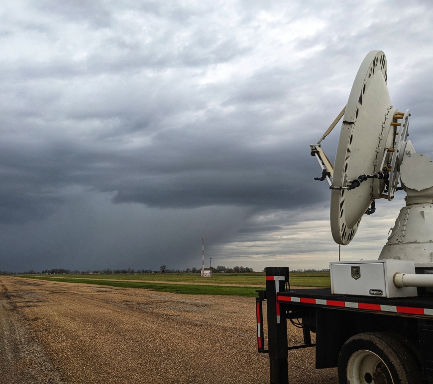 The back bed of a large truck is visible on the right side of the image, with a satellite dish on the bed. It is located on the runway of a small airport. In the background, a dark storm cell is visible, including the frontline clouds and where the rain begins.