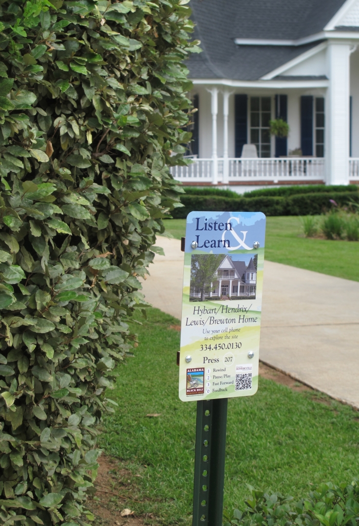 A "Listen & Learn" sign sits in front of the Hybart/Hendrix/Lewis/Brewton Home. The sign offers a phone number to call or QR code to scan to access the audio tour. Behind the sign, a shrub and a section of the house and its front lawn and driveway are visible.