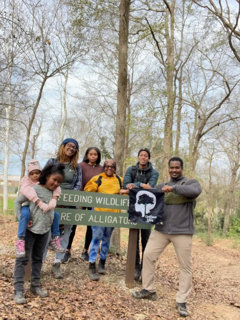 Five adults and two children stand around a trail sign that says "No Feeding Wildlife. Beware of Alligators." They are holding a sign with the Outdoor Afro logo on it.