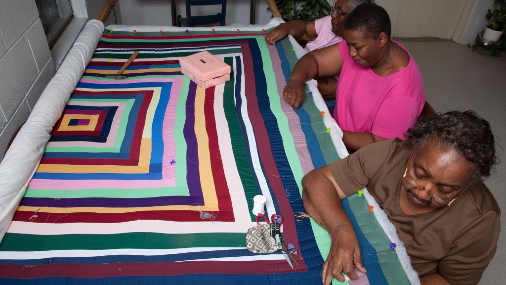Three older women sit in a row on the right side of a quilt, which is partly rolled up. Scissors, thread and other sewing tools are set on the quilt, which displays the bold colors and geometric designs common among Gee's Bend quilts.