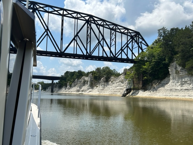 Cliffs with horizontal stripes of white chalk sediment are viewed from a boat, which is only partially visible. Two bridges span the river, one made of metal framing and the other made of concrete.
