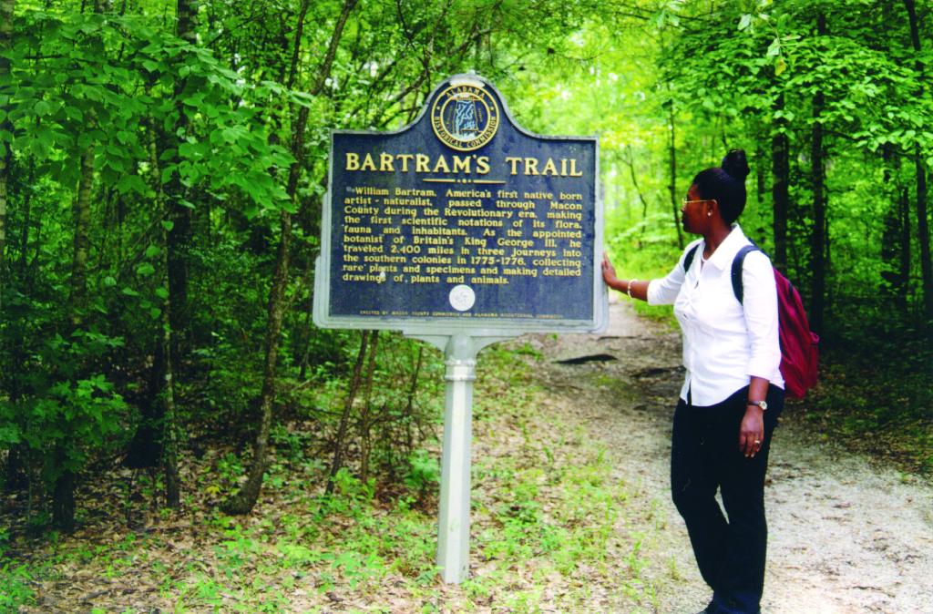 A Black woman with a backpack and dress clothes stands on the hiking trail and reads the sign marking it as a National Recreation Trail. The sign reads: "Bartram's Trail. William Bartram, America's first native born artist-naturalist, passed through Macon County during the Revolutionary era, making the first scientific notations of its flora, fauna and inhabitants. As the appointed botanist of Britain's King George III, he traveled 2,400 miles in three journeys into the southern colonies in 1775-1776, collecting rare plants and specimens and making detailed drawings of plants and animals."