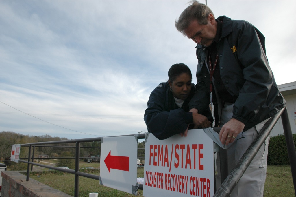 A Black woman and white man tape a sign that reads "FEMA/State Disaster Recovery Center" to a metal fence in front of a building. Other signs with arrows pointing to the center have already been taped up.