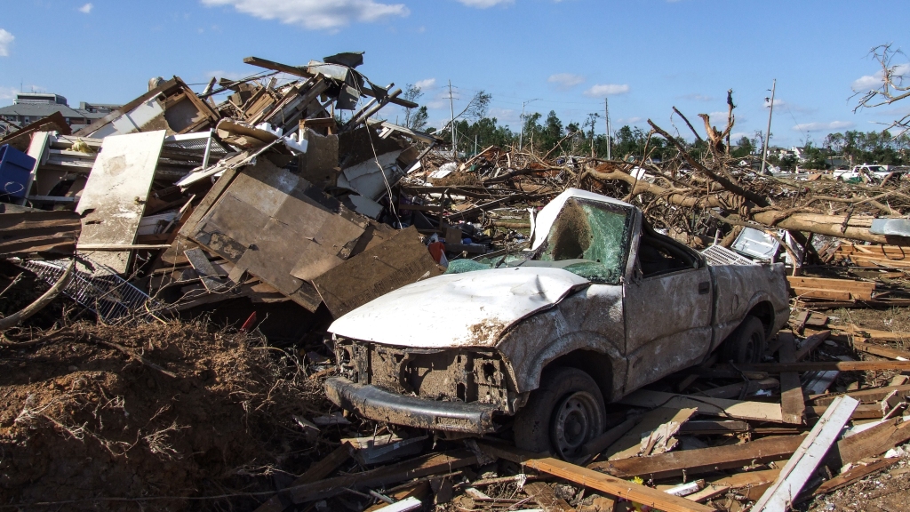 A mostly crushed pickup truck sits on top of piles of boards, with more debris from buildings and trees next to and behind it.