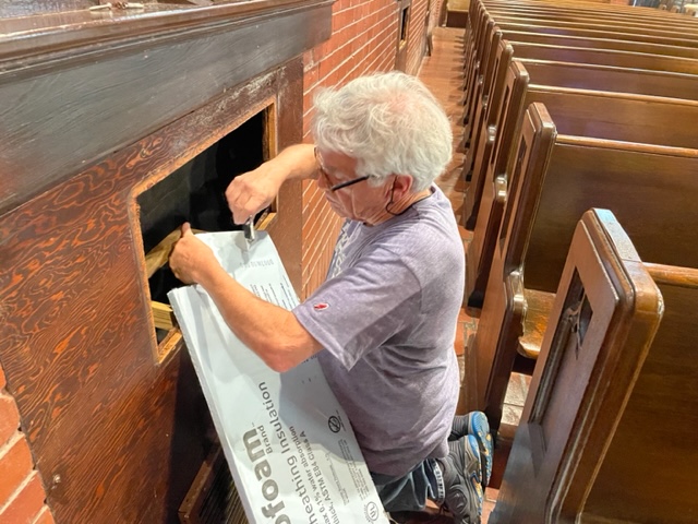 A man cuts open a piece of insulation while kneeling next to an open cut in the wall to reach the ducts. A row of pews are visible behind him.