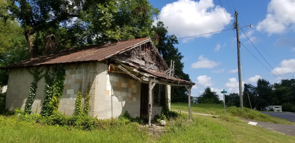A cement-block building in obvious disrepair, with its front awning rotting and grass, vines and kudzu growing around the building and up its walls.