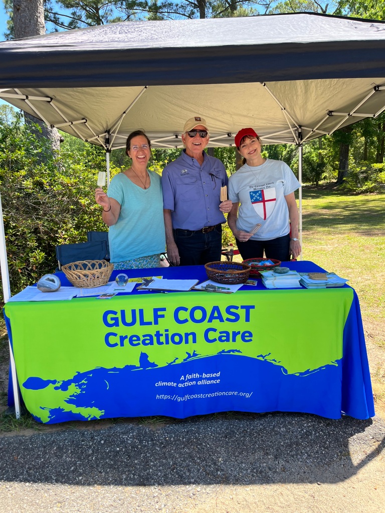 Two women and a man stand under a pop-up tent with a table full of papers and other resources. The table has a banner with Gulf Coast Creation Care's logo on it.