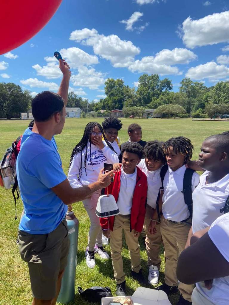 Around 8 middle schoolers stand in an open field listening to an adult, who's holding a weather balloon and a helium tank.