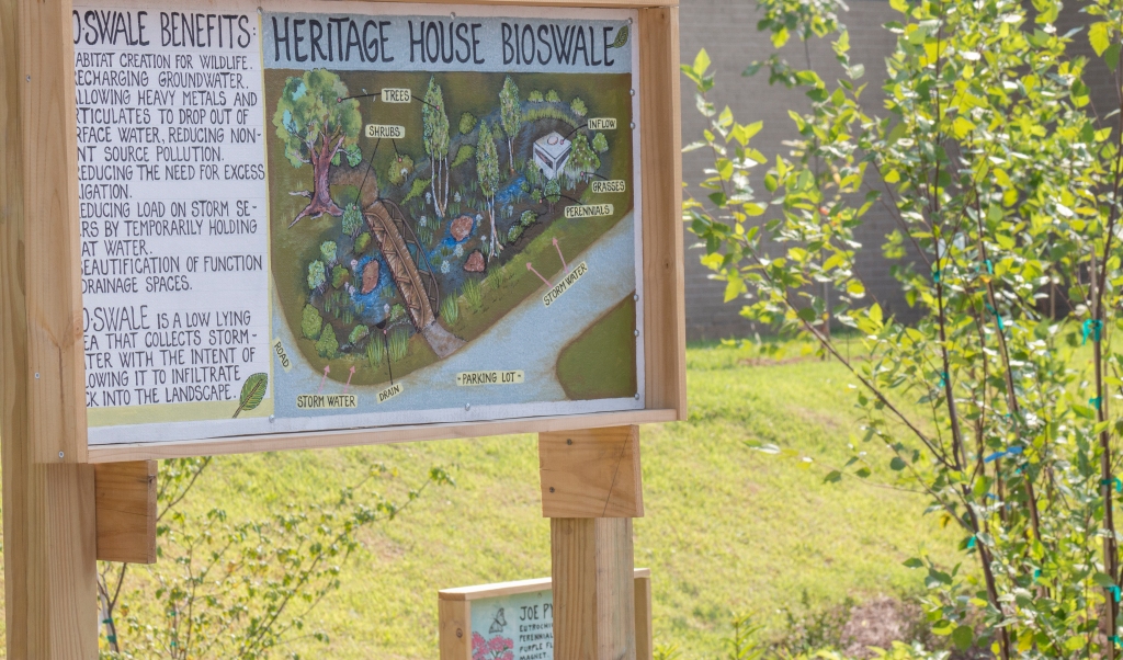 A hand-drawn sign stands next to a sloped grassy area with some plants. The sign includes a drawing of the bioswale, which depicts collected rainwater and several types of plant life, as well as a list of the benefits of a bioswale.
