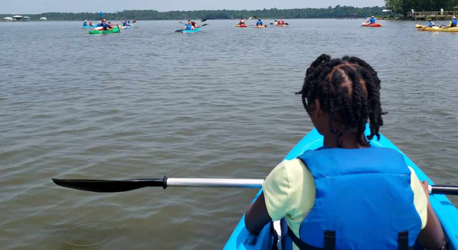 A young Black girl sits on a kayak looking away from the camera, out over the water, with her paddle resting across her lap. She's wearing a life jacket. In the background, several other kayaks are visible.
