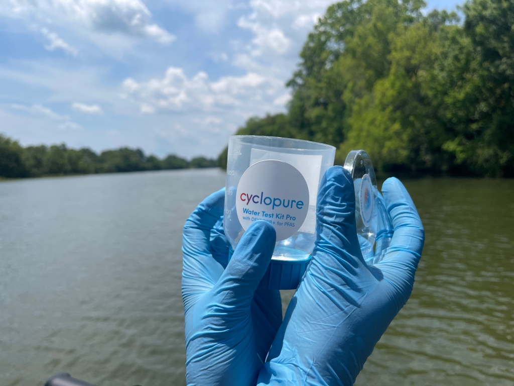 A pair of gloved hands hold a plastic measuring cup and lid, branded "Cyclopure Water Test Kit Pro," in front of a river.
