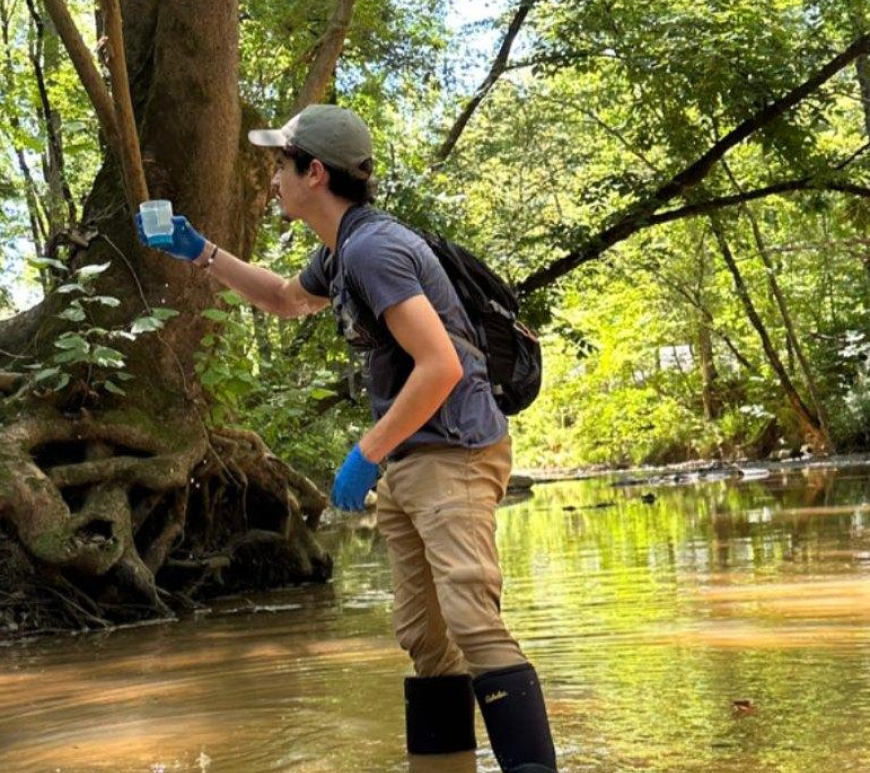 A man wades in a shallow creek with a sampling cup, gloves and a backpack.