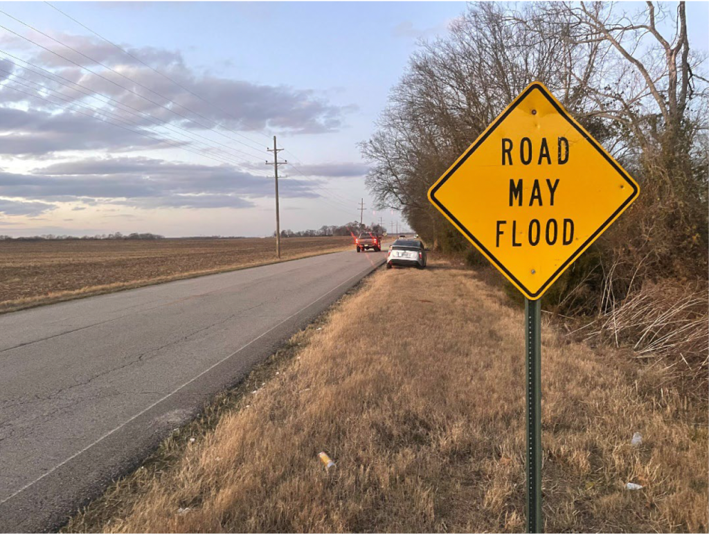 A diamond-shaped sign on the shoulder of a two-lane road reads "Road May Flood."