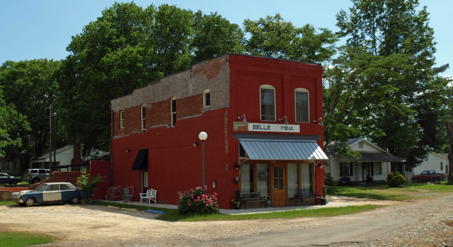 A two-story red building sits in front of a broken asphalt road with a gravel parking lot next to it. The building appears to have been part of a row of shops at one point, which are no longer there. A few houses are visible behind it. Over the front door awning, a sign reads "Belle Mina."